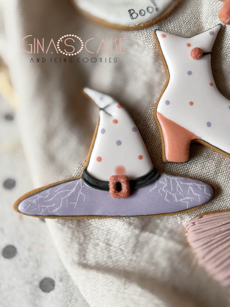 Due biscotti decorati a tema Halloween: un cappello da strega con dettagli viola e un paio di stivali rossi.