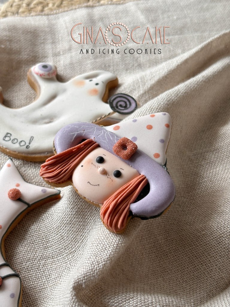 Biscotti decorati a tema Halloween, tra cui un volto di streghetta con cappello viola e capelli rossi, su uno sfondo di tessuto beige.