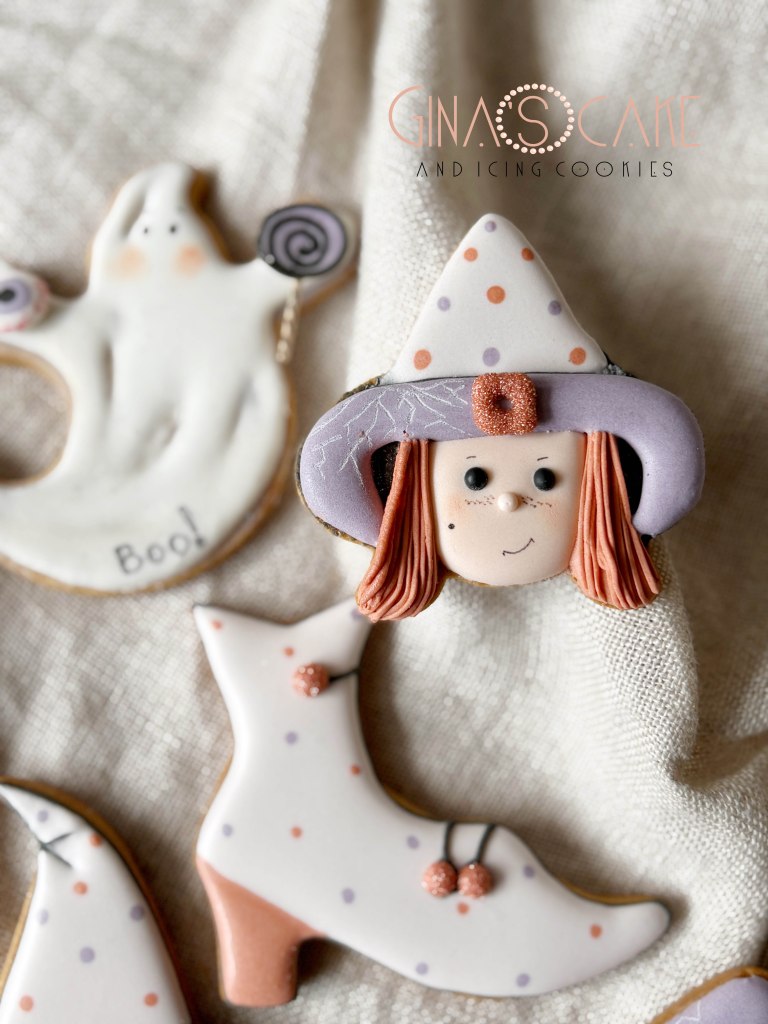 Biscotti decorati a tema Halloween, tra cui un fantasmagorico fantasmino, un cappello di strega con una faccia sorridente e stivali bianchi.