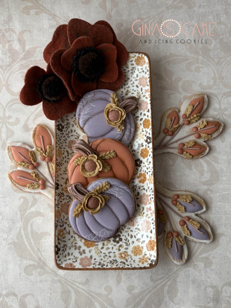 Un vassoio decorato con biscotti a forma di zucca in tonalità viola e arancione, adornati con dettagli floreali, accompagnati da fiori in feltro rossi e decorazioni autunnali.
