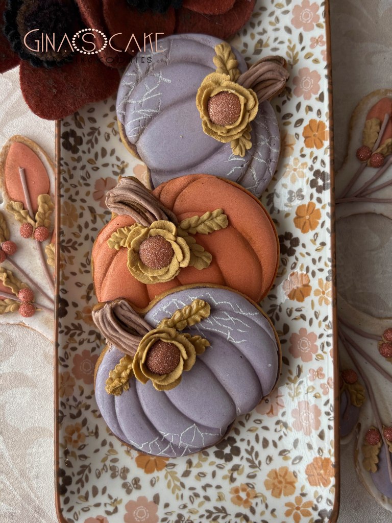 Tre biscotti a forma di zucca decorate con fiori in pasta di zucchero, su un piatto ornamentale.