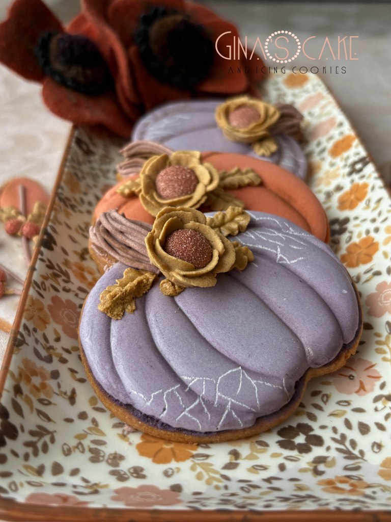 Tre biscotti decorati a forma di zucca in diverse tonalità, con fiori in pasta di zucchero e dettagli ornamentali su un piatto decorativo.