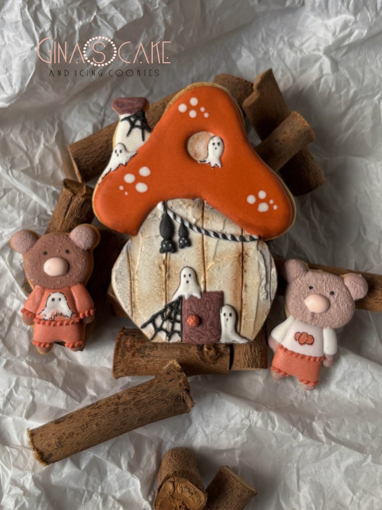 Due orsetti di biscotto con una casa a forma di fungo decorata con fantasmi, circondati da bastoncini di legno.