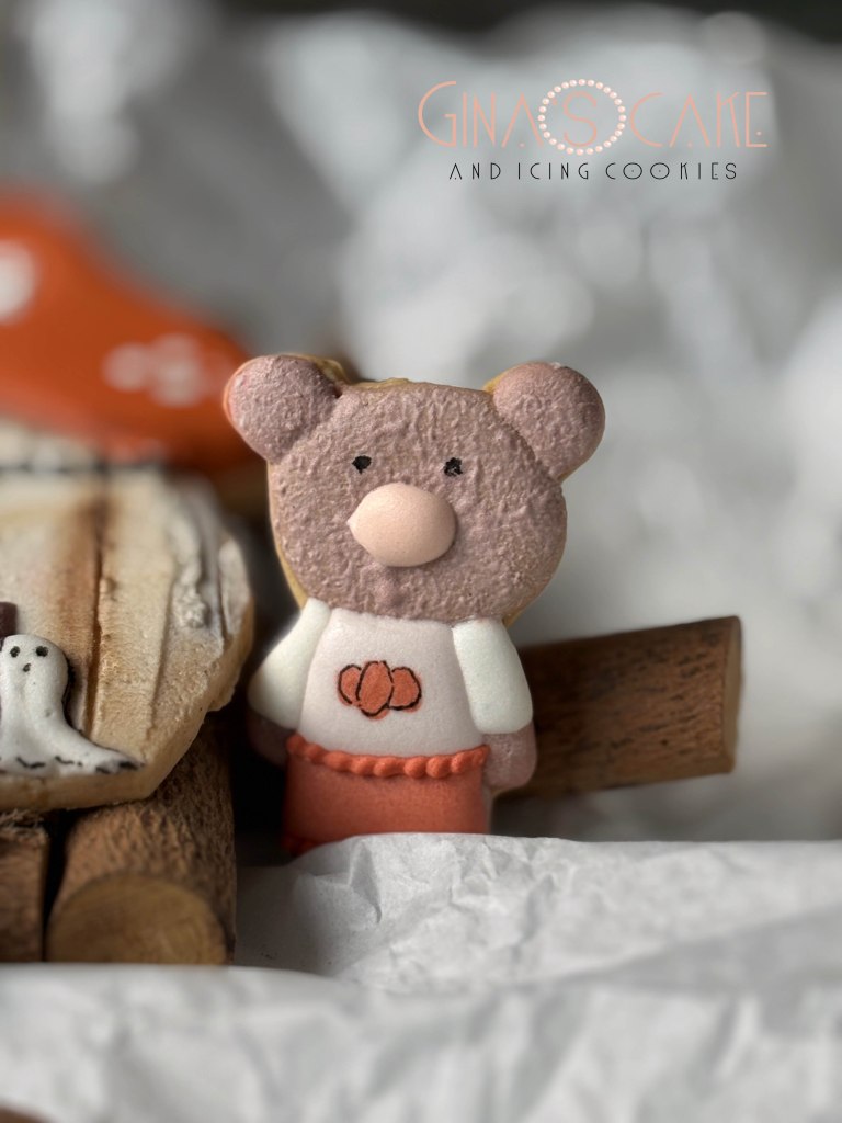 Un portachiavi a forma di orsetto di pasta di zucchero, con una maglietta bianca e una gonna arancione, posizionato su un fondo di carta e rami decorativi.