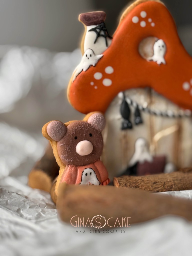 Un biscotto a forma di orsetto carino con un maglione arancione e un fantasma, posato davanti a una casetta decorata per Halloween.